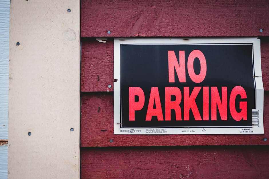 Close-up view of a rectangular parking restriction sign mounted on a metal pole against a backdrop of leafy green trees and a partly cloudy sky. The sign features a bold red circle with a diagonal lin