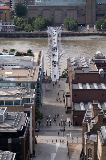 A view of a large brick building with four tall chimneys, situated along the riverbank near Tate Britain in Pimlico. Adjacent to the building, there are modern glass structures reflecting the cloudy s