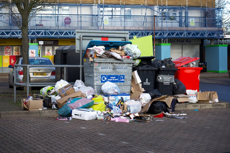 A large, green waste or recycling container positioned on the pavement outside a modern building with a grey tiled facade and large glass windows. The container features multiple metal latches and doo