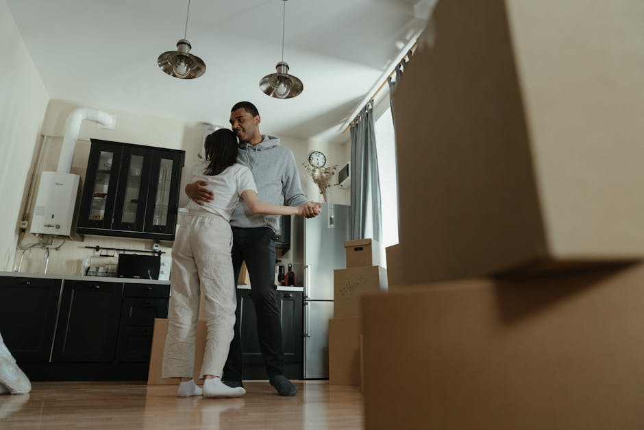 Inside a residential property during a home relocation, a man and a woman are embracing each other in the kitchen area, which features dark lower cabinets, a wall-mounted black glass-fronted cabinet, and a white boiler. The woman is dressed in light-colored casual clothing, and the man is wearing a grey hoodie and dark pants. The kitchen is well-lit by natural daylight streaming through a window with light blue curtains, and there is a small wall clock and a decorative plant on a shelf in the background. To the right, several cardboard moving boxes are stacked on the wooden floor, indicating packing activities. The scene captures the loading process involved in furniture transport and house removals, with the focus on emotional moments during a household move, supported by professional relocation services like those offered by Pimlico Man and Van.
